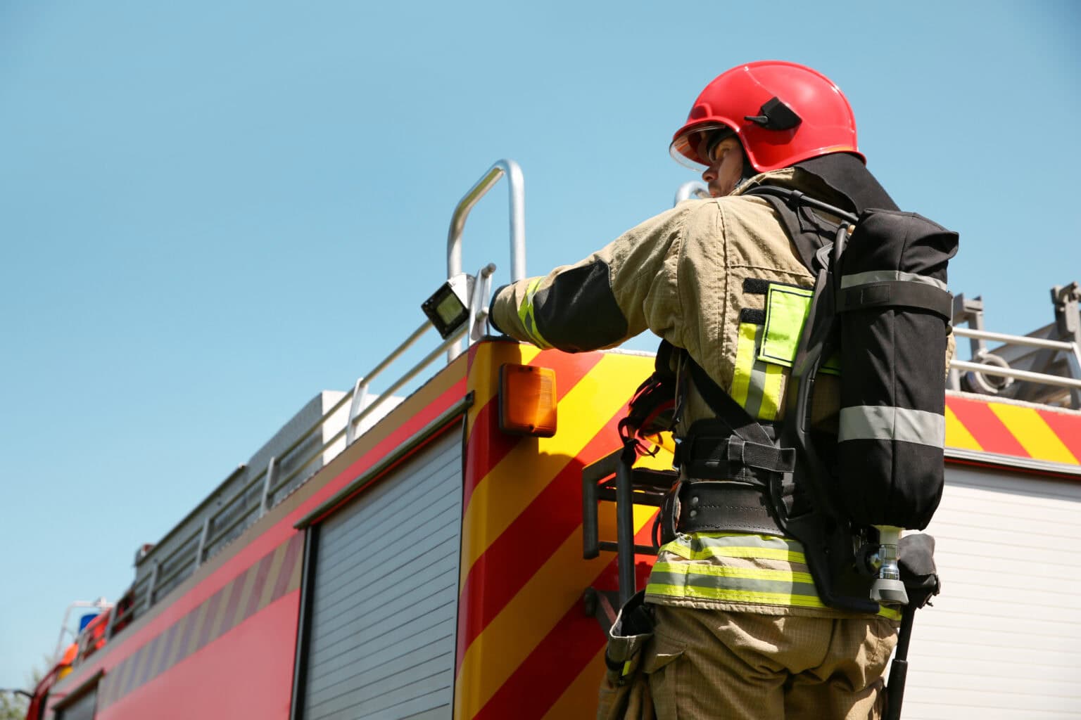 Firefighter in gear preparing on a fire truck under a clear blue sky.